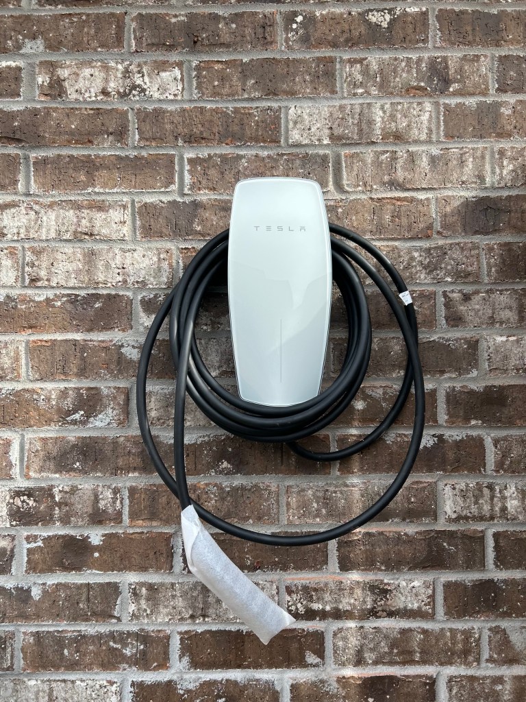 A Tesla wall-mounted electric vehicle charger with cables hanging down, installed on a brick wall.