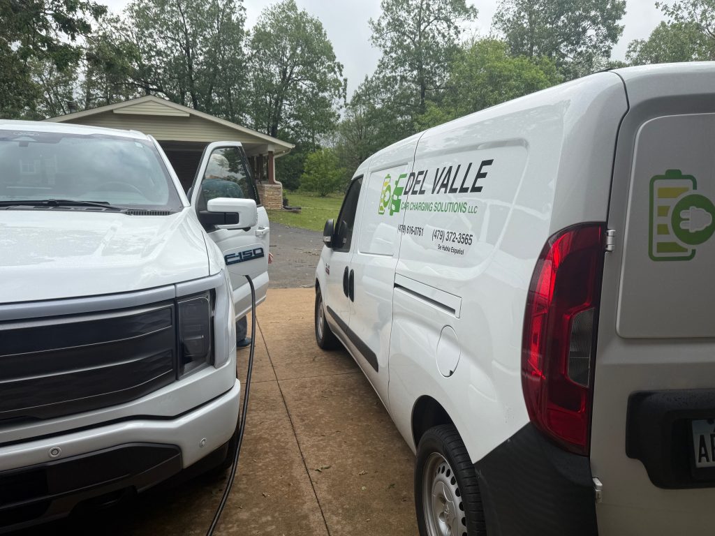 A white electric vehicle and a service van from Del Valle Car Charging Solutions parked in a driveway, showcasing EV charging equipment.